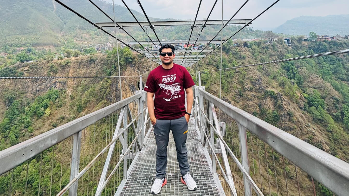 A man stands confidently in the middle of a suspension bridge spanning a deep gorge, surrounded by green hills and distant buildings.