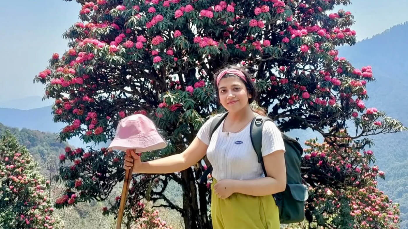 A woman stands in front of a blooming pink rhododendron tree during a hike, holding a walking stick with a pink hat and wearing a backpack.
